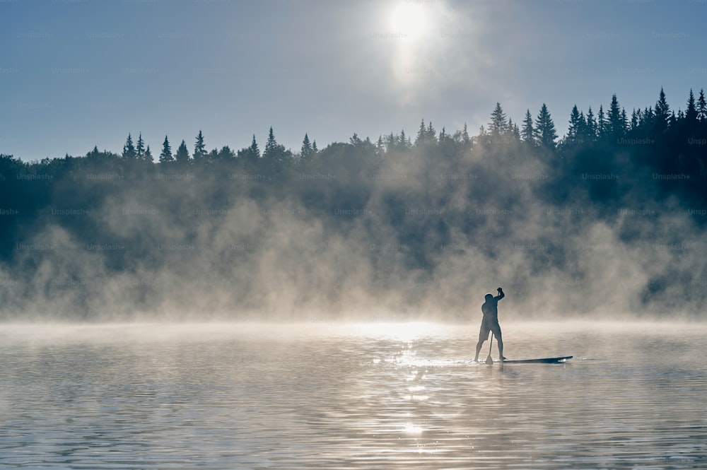 Silhouette of a man paddling on surf board in early morning when fog is rising in the mountains