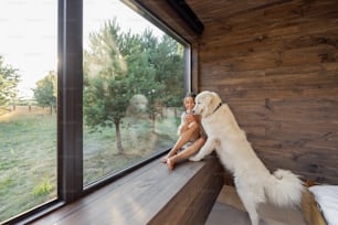 Young woman resting at beautiful country house or hotel, sitting on the window sill with pine forest view and hugs with big white dog. Concept of solitude and recreation on nature with pet