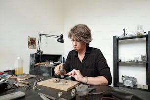 Busy middle-aged woman in black shirt sitting at desk in workshop and soldering Jewelry while finishing work