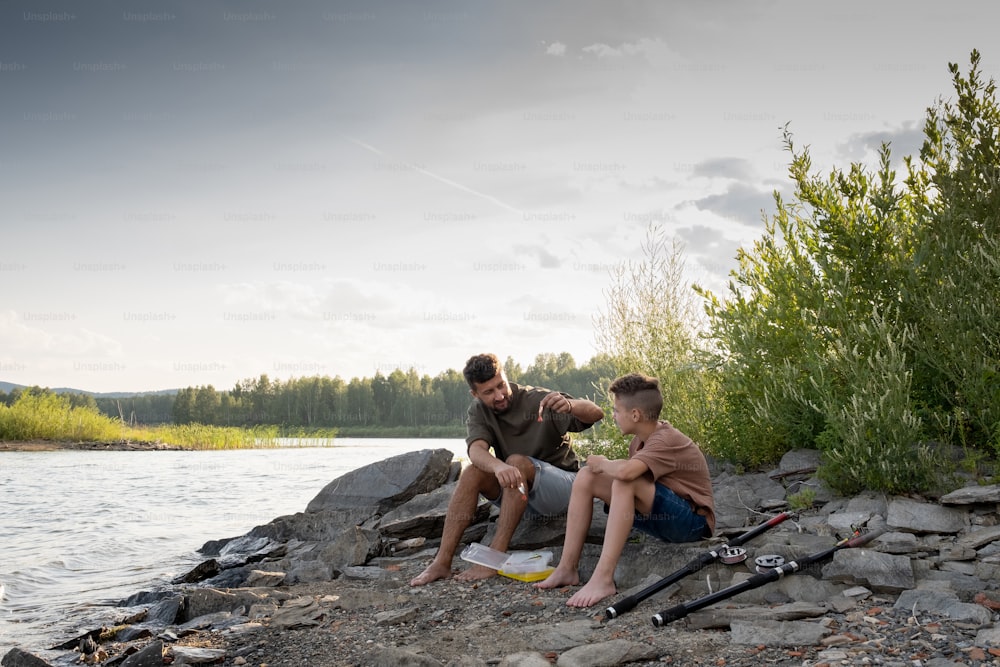 Father and son sitting in front of lake while fishing on summer weekend