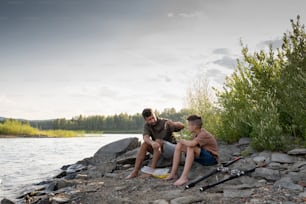 Father and son sitting in front of lake while fishing on summer weekend