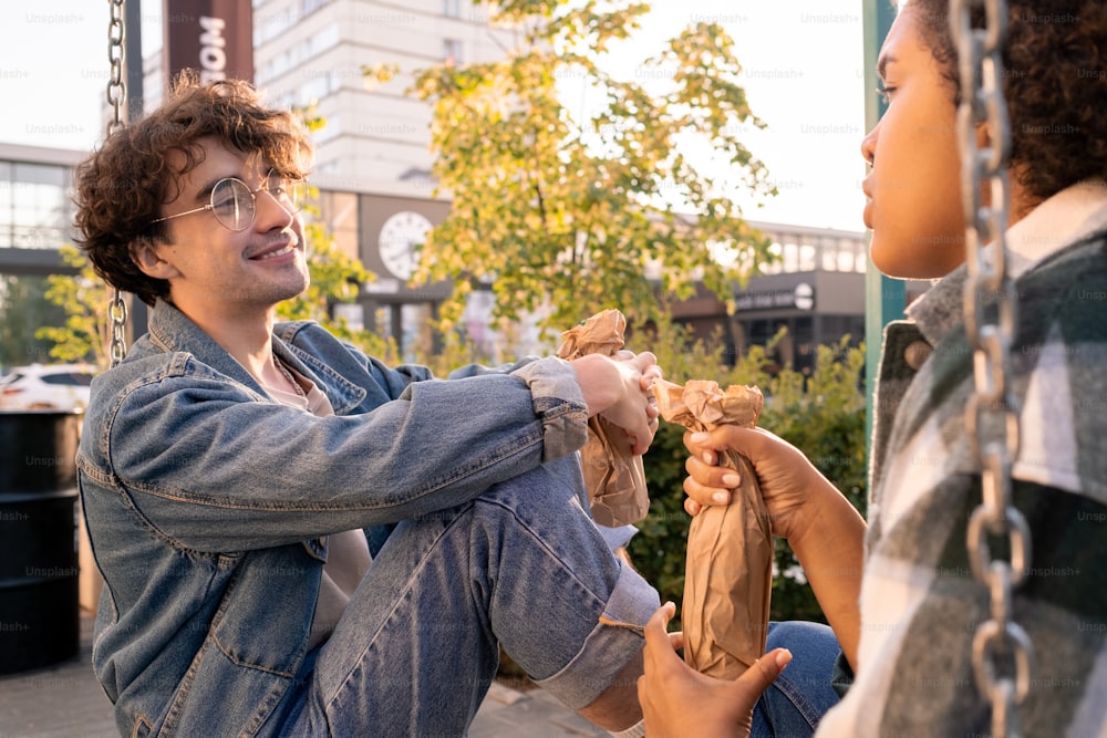Smiling teenager with bottle looking at his girlfriend during outdoor discussion