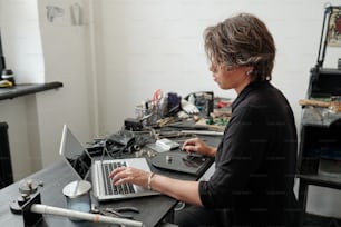Middle-aged jeweler sitting at desk with tools and using laptop while creating content for online jewelry shop