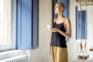 Young stylish woman stands with a coffee near the window at modern bright apartment. Comfort living and leisure time at home