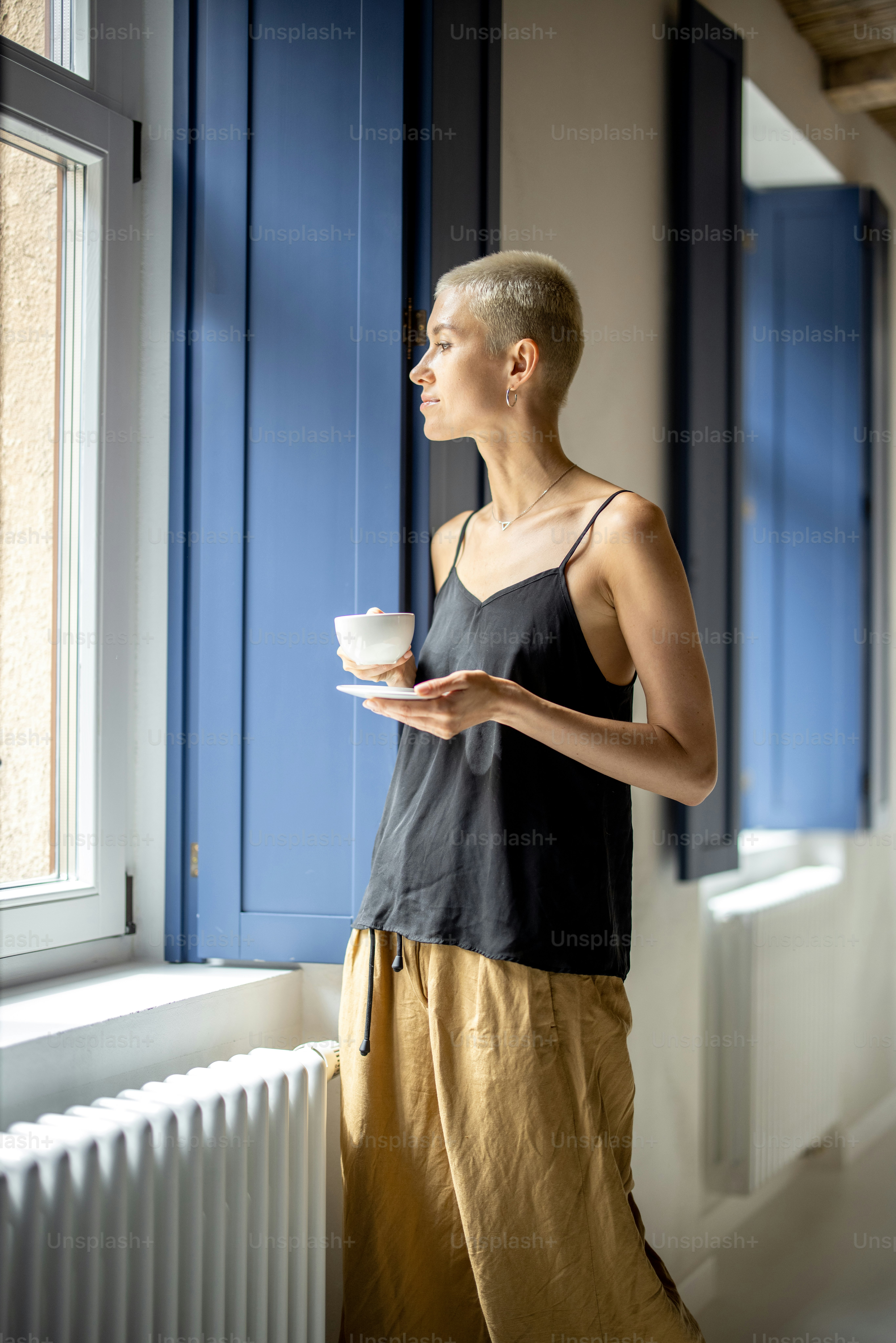 Jovem mulher elegante fica com um café perto da janela do apartamento moderno e brilhante. Vida confortável e tempo de lazer em casa