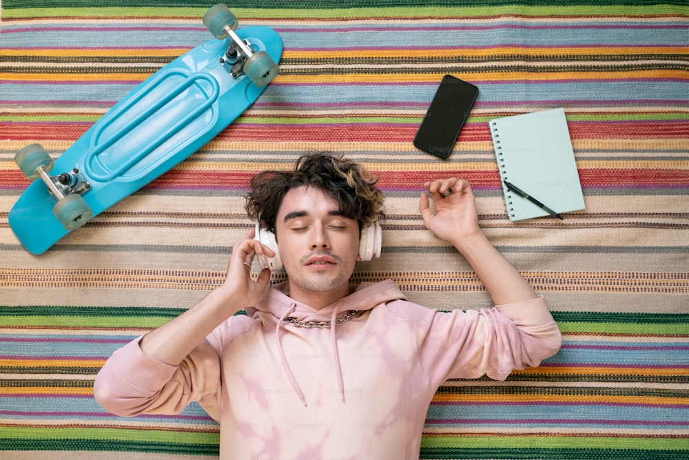 Restful guy in headphones and pink hoodie listening to music on the floor among skateboard, notepad and smartphone