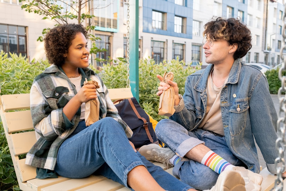 Young intercultural couple in casualwear having drinks while relaxing on swings outdoors