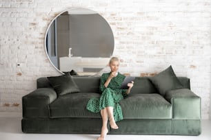 Stylish woman in green dress using digital tablet while sitting relaxed on the comfortable sofa at home. Leisure time with a digital device or working home concept