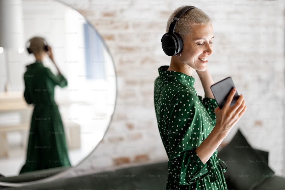 La mujer elegante con vestido verde disfruta de la música con auriculares y teléfono celular bailando en la sala de estar de casa