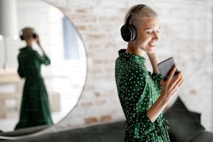 Stylish woman in green dress enjoys the music with headphones and cell phone dancing in the living room at home