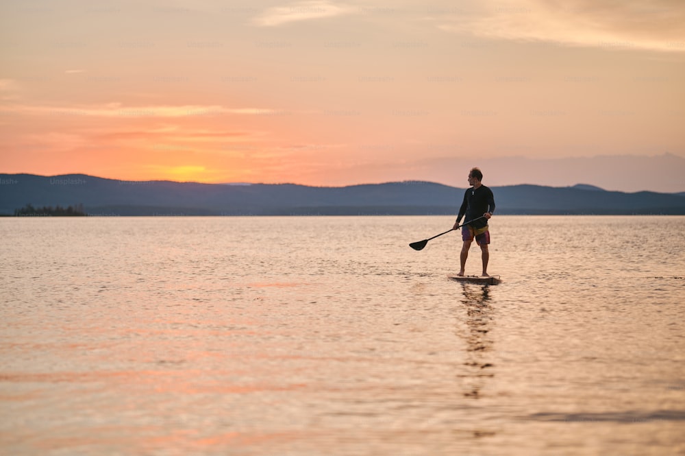 Full body shot of a man with paddle standing on wooden board in quiet water, surfing on sunset with scenic mountain view