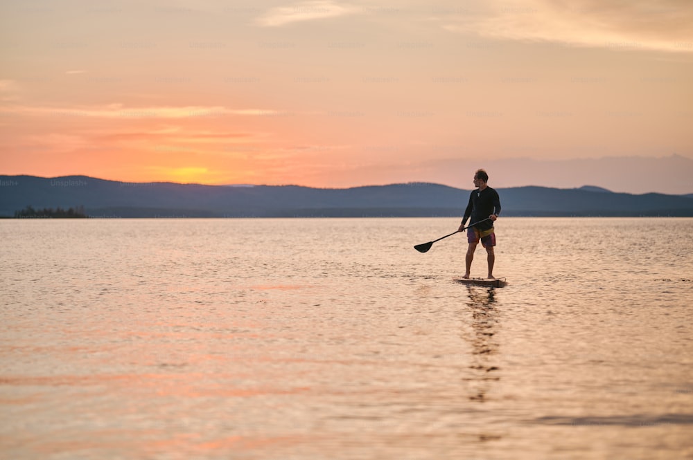 Ganzkörperaufnahme eines Mannes mit Paddel, der auf Holzbrett in ruhigem Wasser steht und bei Sonnenuntergang mit malerischem Bergblick surft