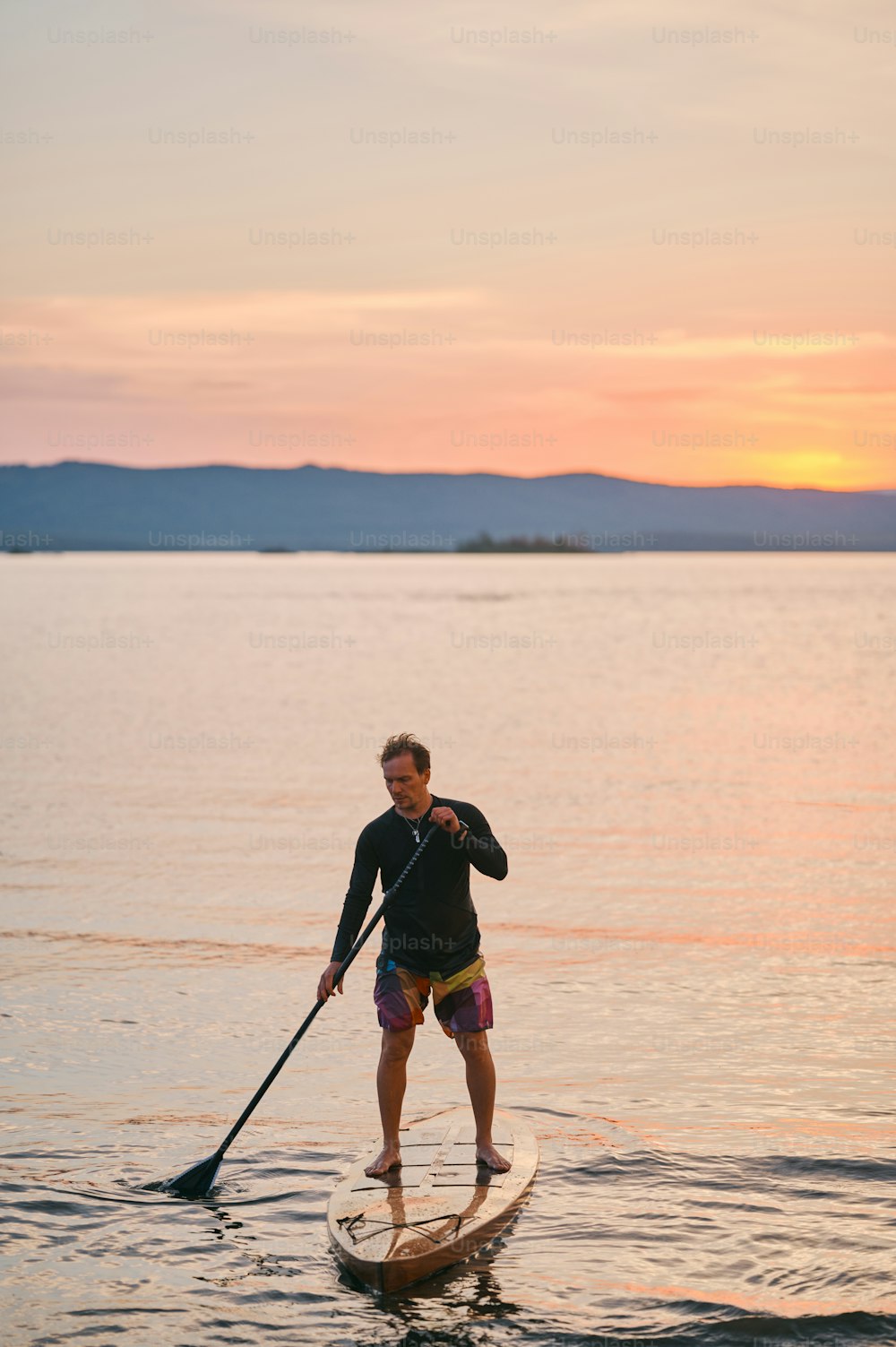 Full body shot of a man with paddle standing on wooden board in quiet water, surfing on sunset with scenic mountain view