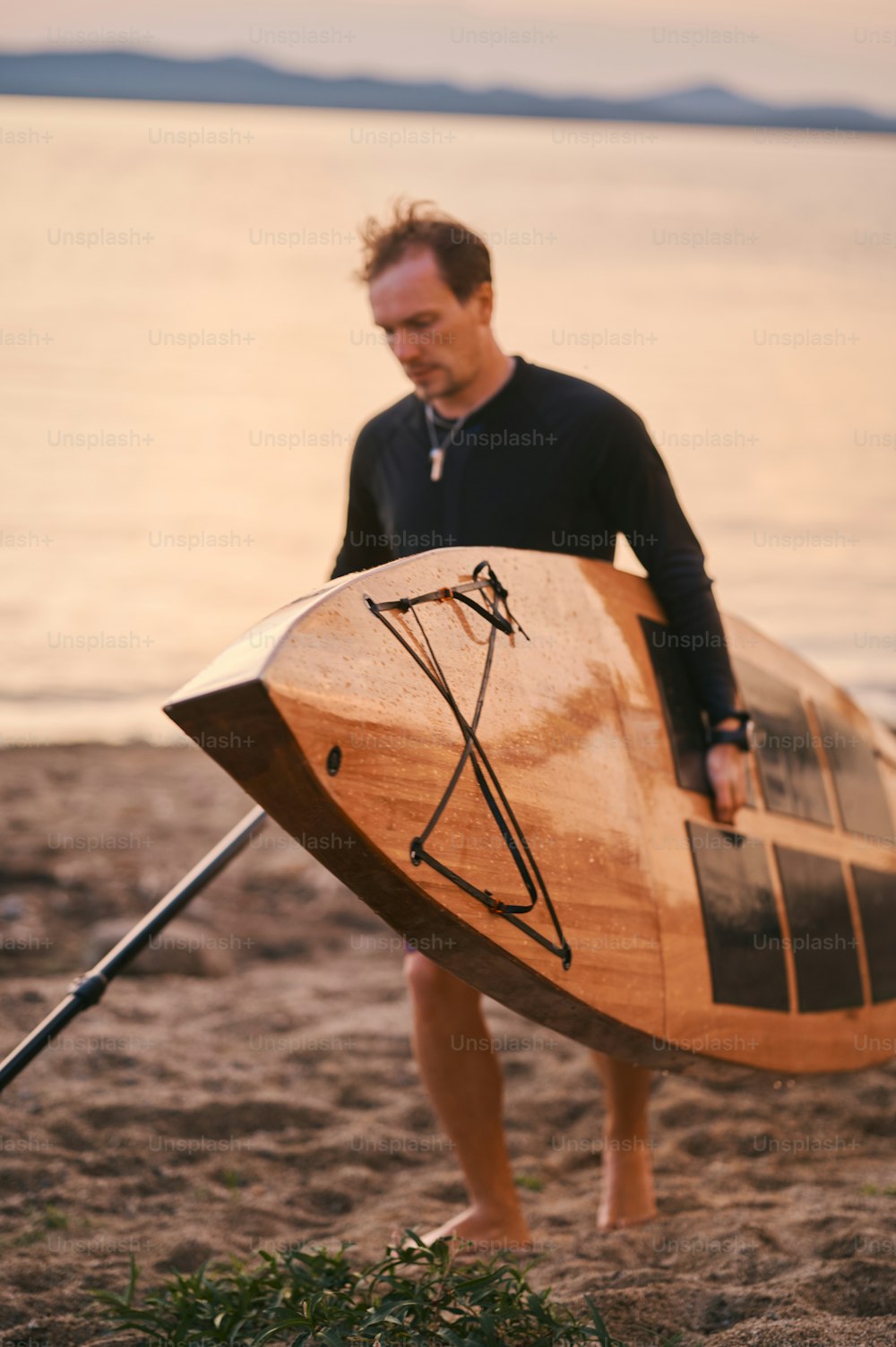 Foto de corpo inteiro de um homem forte carregando prancha de surf e um remo, saindo da água depois de nadar