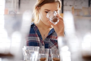Portrait in selective focus of young female barista examining cups with beans and ground coffee, doing cupping test, smelling fresh coffee