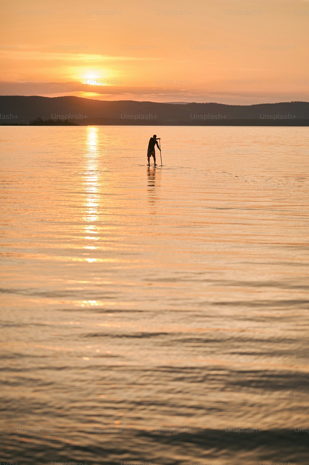 Figure d’un homme debout sur une planche de sup surf, pagayant sur une eau calme au coucher du soleil