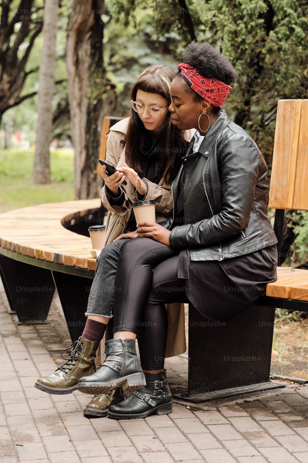 Two casual girls discussing photos in smartphone while relaxing on bench in public park