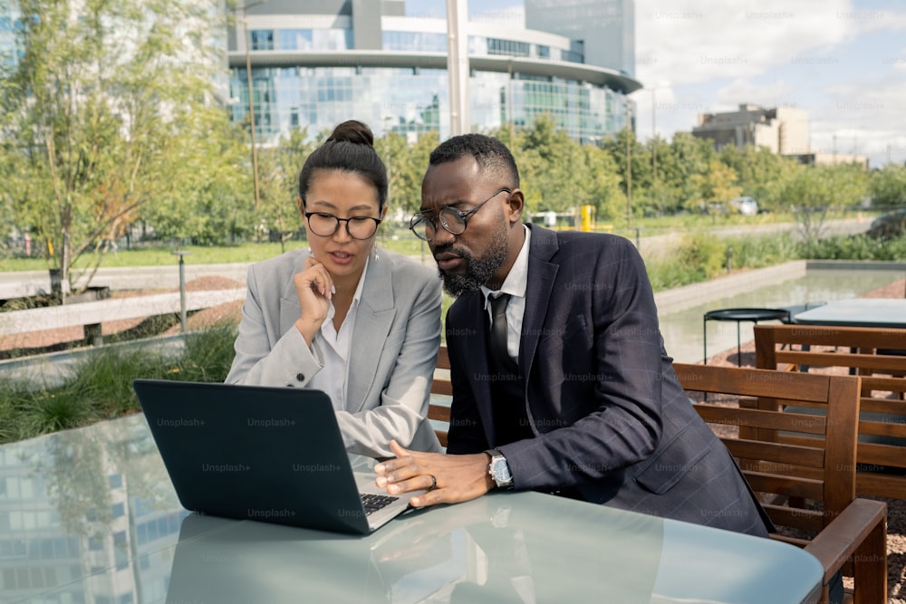 Two young colleagues in formalwear and eyeglasses sitting by table in front of laptop outdoors