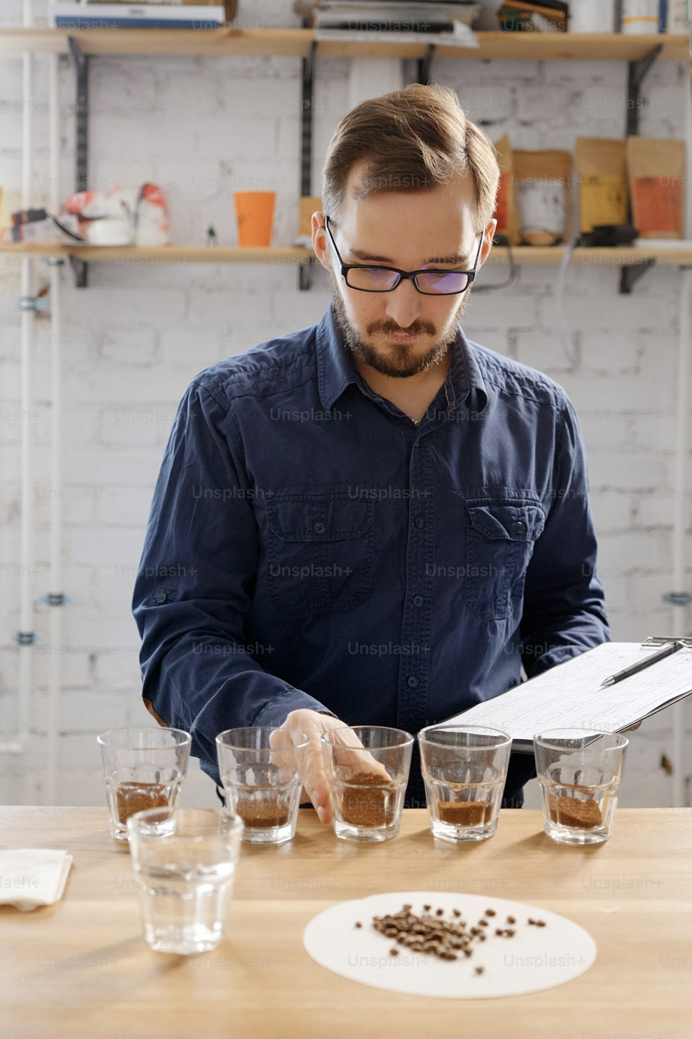 Portrait of man smelling freashly ground coffee in cups at coffee cupping examine. He is standing near white wall in front of rows with glass cups