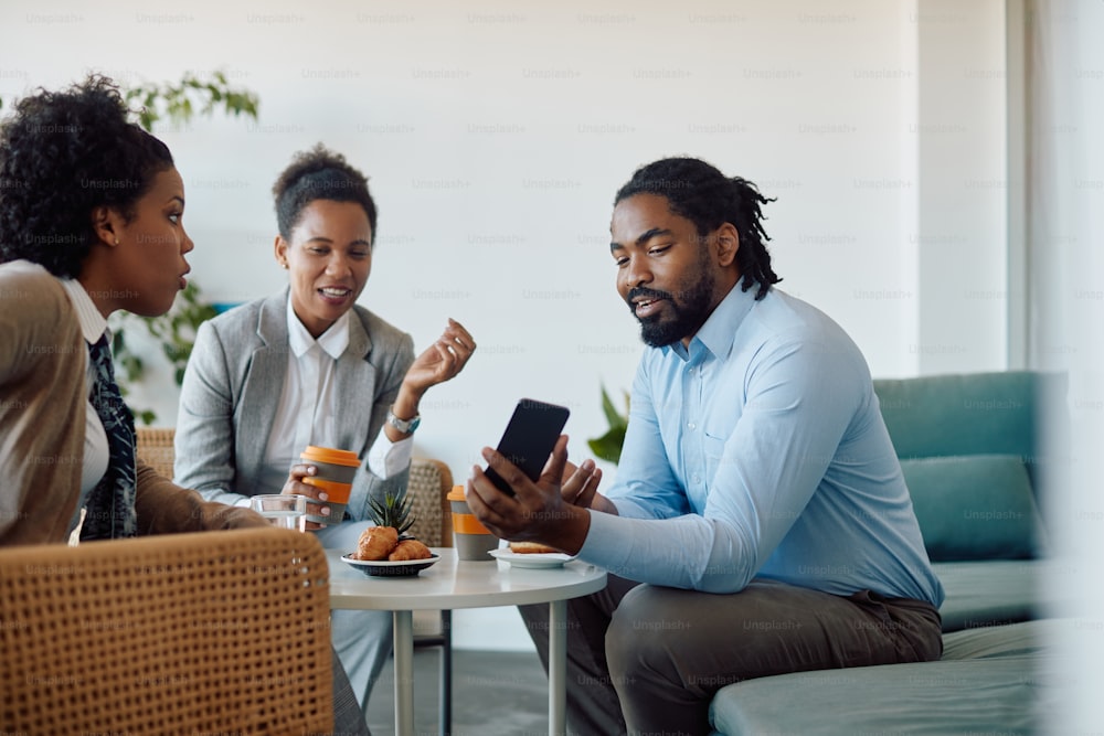 African American businessman showing something on mobile phone to female colleagues during coffee break at work.