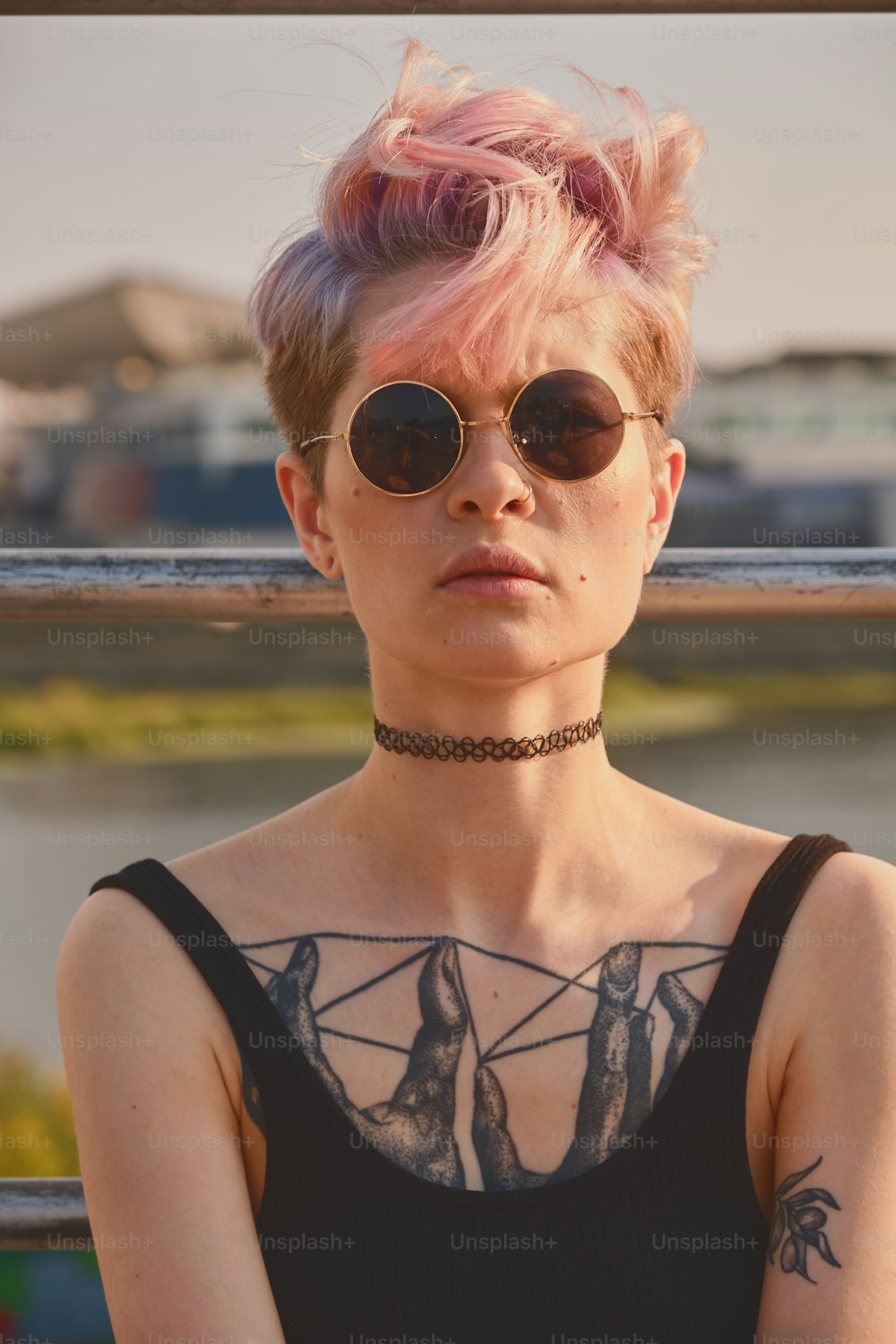 Retrato da mulher tatuada jovem ousada com cabelo rosa curto, peito aberto, sentado ao ar livre em óculos redondos