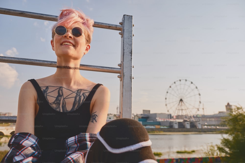 Retrato da mulher tatuada jovem ousada com cabelo rosa curto, peito aberto, sentado ao ar livre em óculos redondos