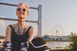 Portrait of daring young tattooed woman with short pink hair, open chest, sitting outdoor in round glasses