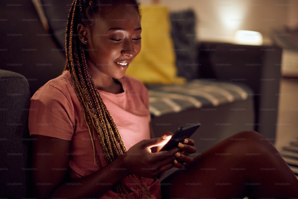 Joven mujer afroamericana usando un teléfono inteligente mientras se relaja por la noche en casa.