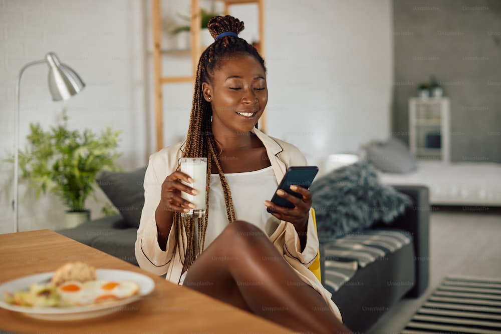 Smiling black woman having a glass of milk and using smart phone during her breakfast at home.