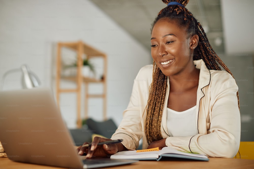 Happy black female student in pajamas e-leaning over laptop at home.