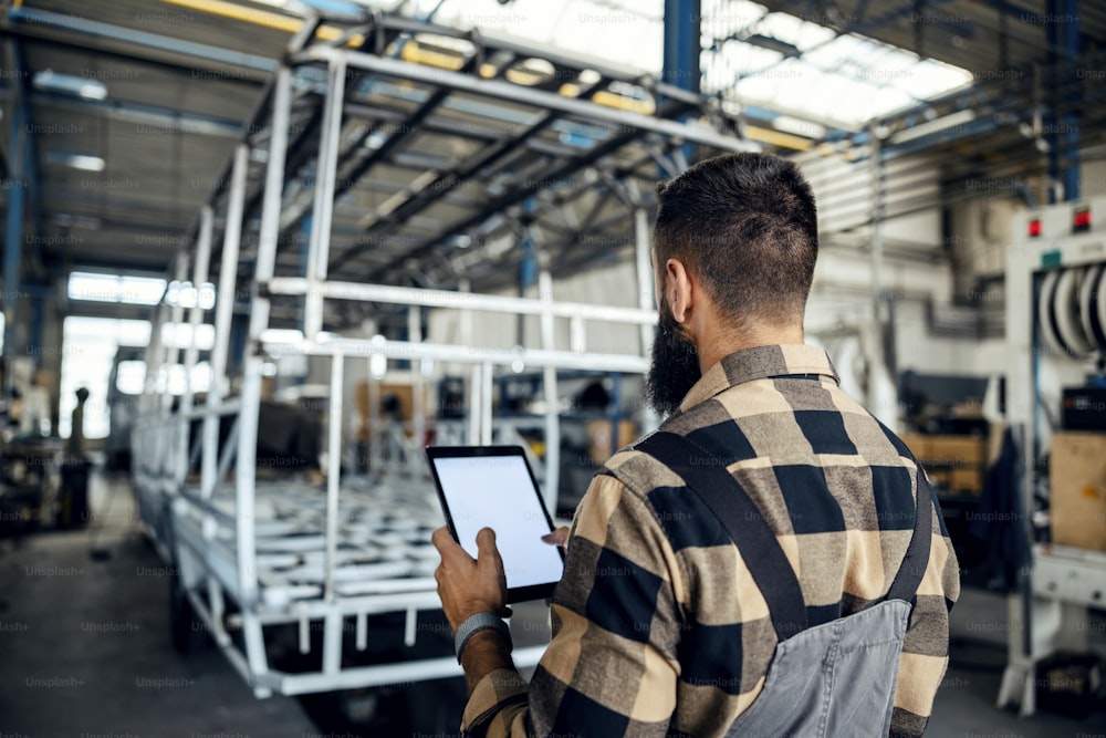 Rear view of auto industry worker standing in a factory with tablet in his hands and looking at metal construction prepared for a new bus. Auto-mechanic in bus factory