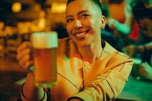 Young happy black woman toasting while drinking beer at night in a pub.