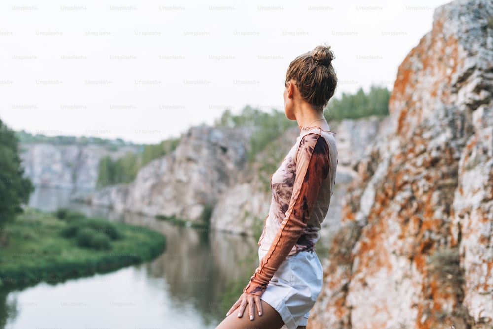 Junge schlanke Frau in Freizeitkleidung blickt auf schöne Aussicht auf Berge und ruhigen Fluss, lokale Reisen