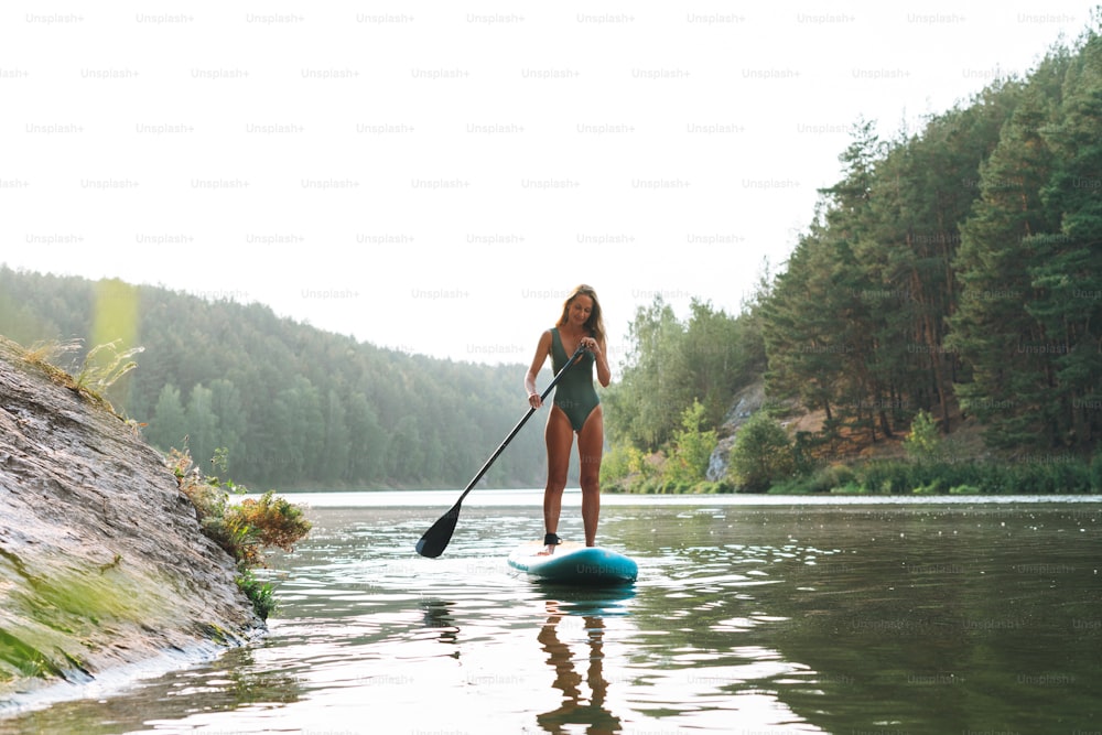 緑色のスウィームスーツを着たスリムな若い女性が、川に浮かぶオールを漕ぐサップボートに乗っている、週末旅行や地方旅行