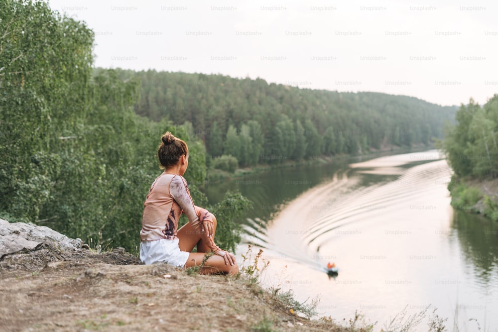 Junge schlanke Frau in Freizeitkleidung blickt auf schöne Aussicht auf Berge und ruhigen Fluss, lokale Reisen