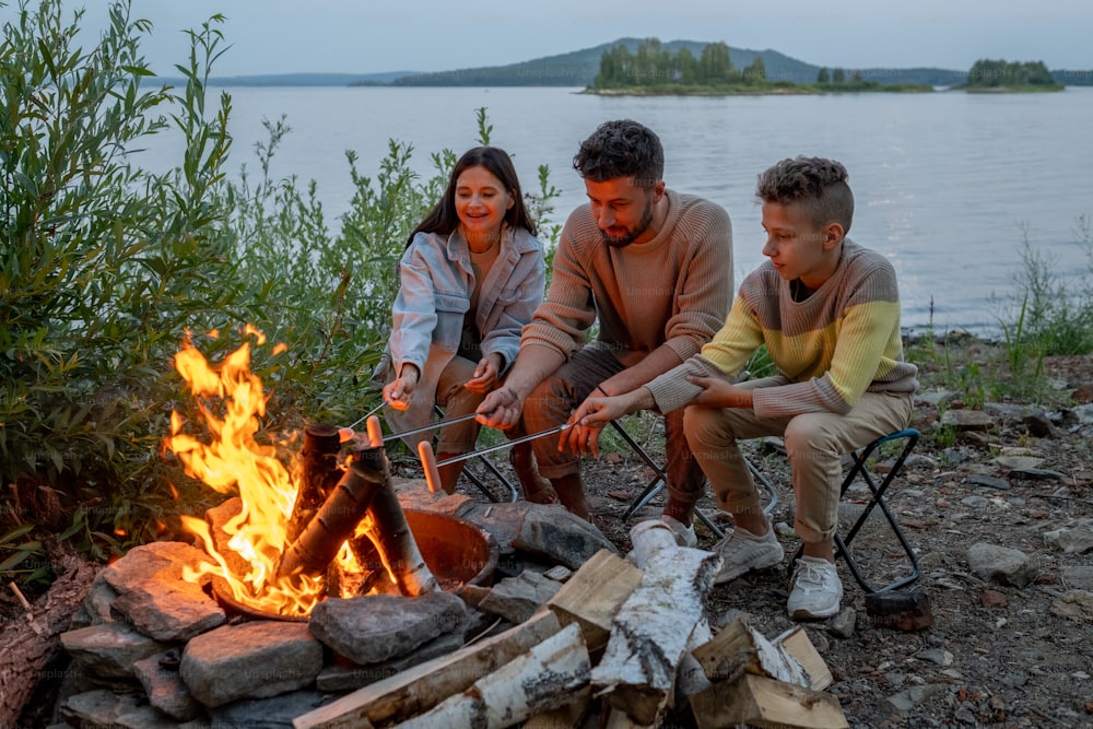 Família de jovens pais e seu filho fritando salsichas de fogueira contra a beira-mar