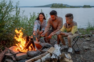 Family of young parents an their son frying sausages by campfire against waterside
