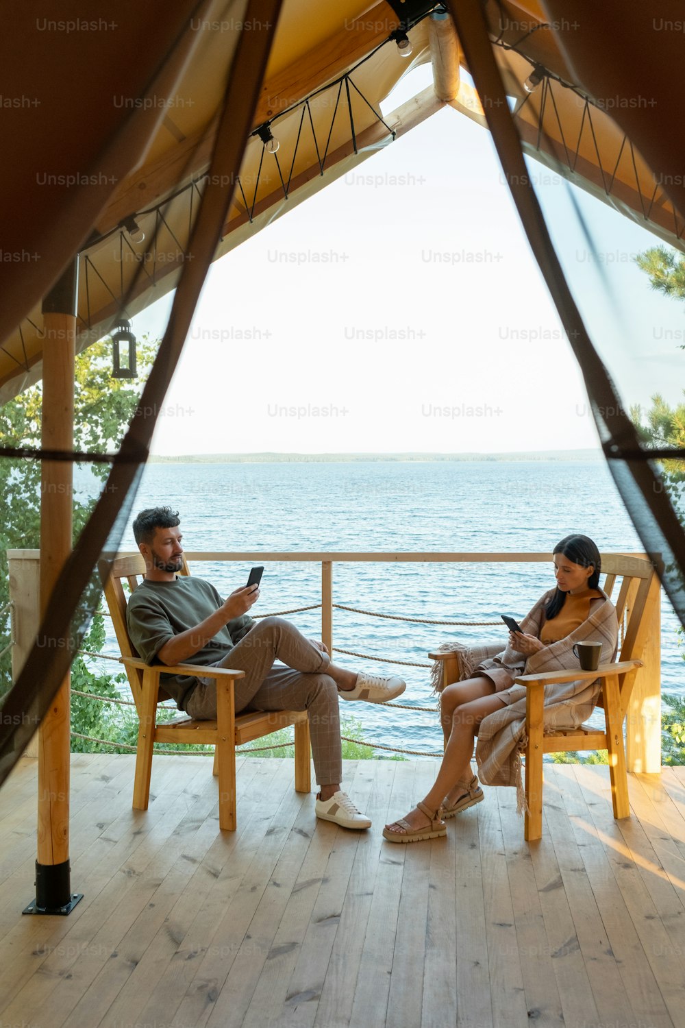 Joven pareja tranquila desplazándose en smrtphones mientras está sentada en sillones de madera contra la orilla del mar