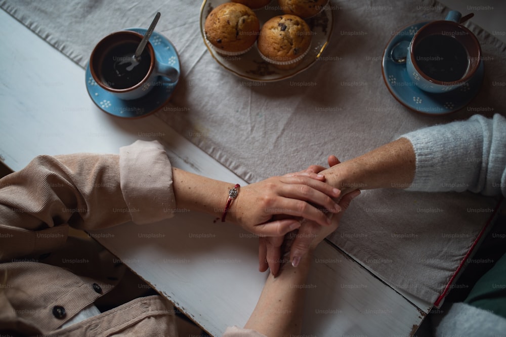 Une vue de dessus d’une mère âgée prenant un café avec sa fille adulte à l’intérieur à la maison, se tenant la main.