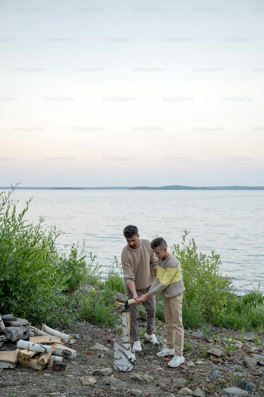 Adolescent boy helping his father to chop firewood while both holding axe over log
