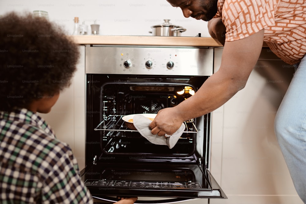 Giovane uomo africano che prende la torta di mele al forno calda dal forno elettrico davanti al suo piccolo figlio