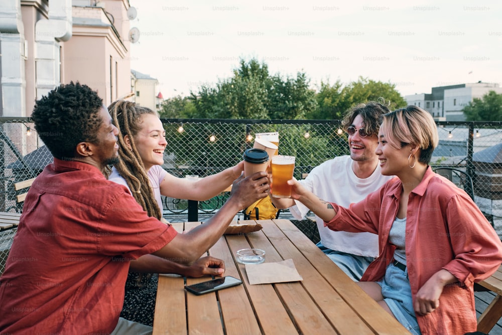 Young cheerful intercultural friends toasting with drinks over wooden table