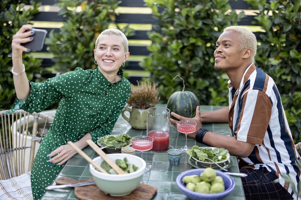 Multiracial couple taking selfie on mobile phone during dinner outdoors. Concept of relationship. Modern lifestyle. Black man and european girl sit at table with organic food. Idea of healthy eating