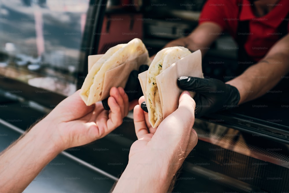 Hände eines jungen Mannes, der leckere Snacks in Papier einwickelt, die von einem behandschuhten Arbeiter eines Imbisswagens gehalten werden