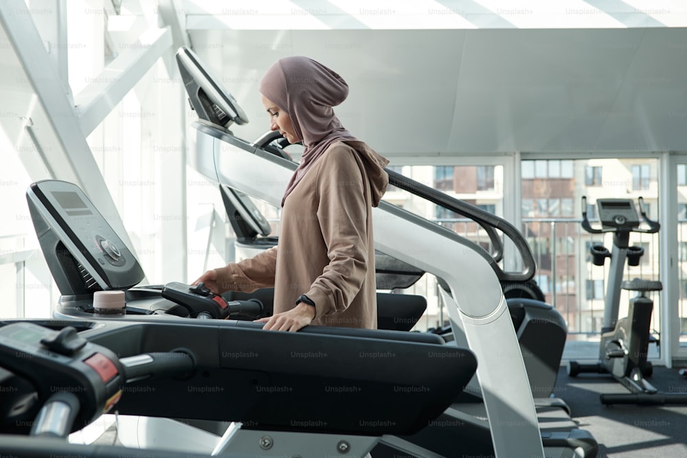 Seitenaufnahme einer schönen muslimischen Frau mit Hijab, die Zeit im Fitnessstudio auf dem Laufband verbringt und die Einstellungen anpasst