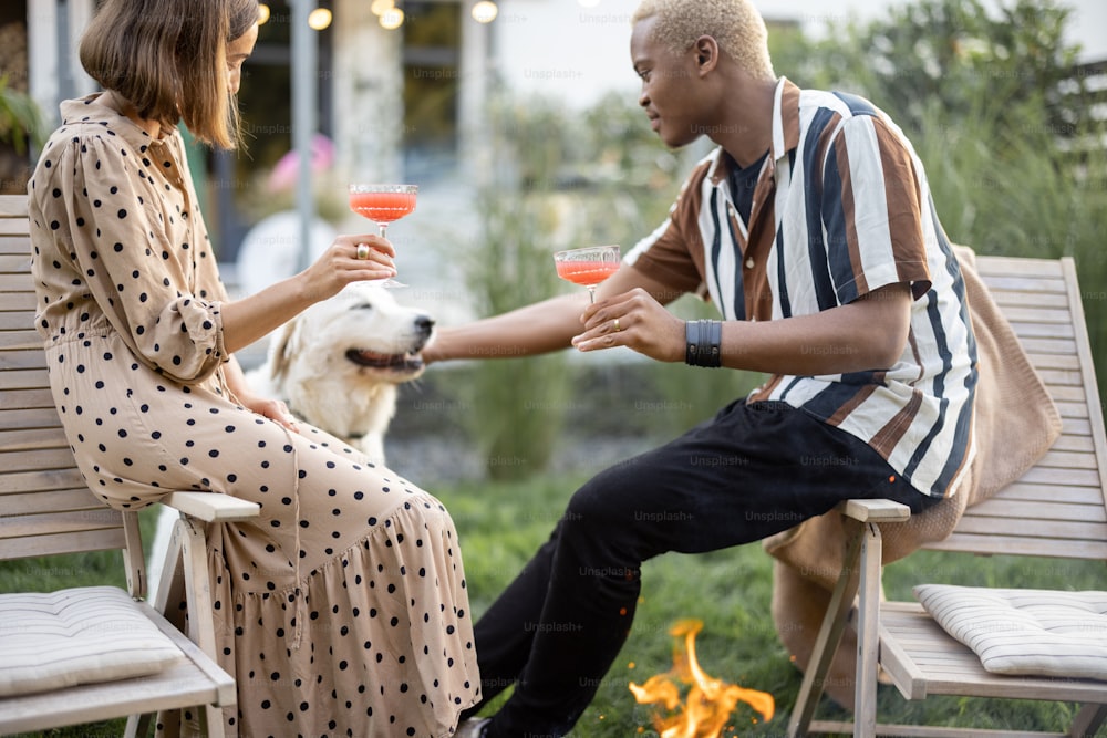 Multiracial couple drinking cocktails and caressing dog during home party in their garden. Burning fire for bbq. Concept of relationship rest. Black guy and european girl enjoying time together.