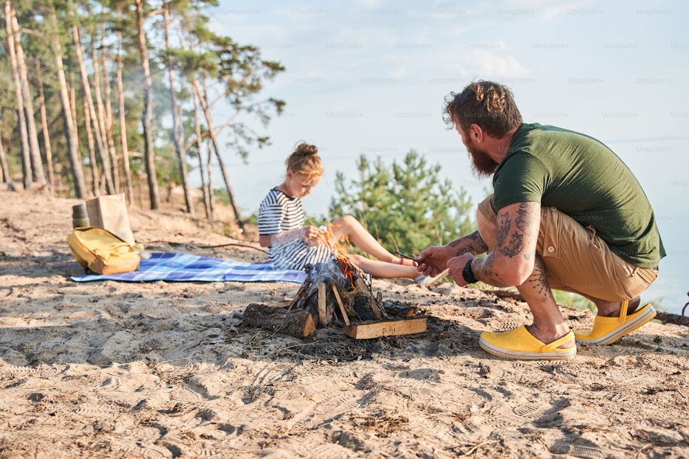 Full length view of the positive relaxed family feeling comfortable together while spending time at the nature. Man preparing campfire while his daughter sitting at the blanket
