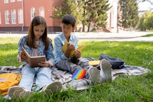 Two cute intercultural schoolchildren reading books while resting on green lawn