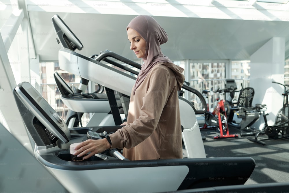 Vista laterale colpo medio di giovane donna musulmana adulta che indossa un copricapo che inizia l'esercizio in palestra in piedi sul tapis roulant che regola le impostazioni prima di correre