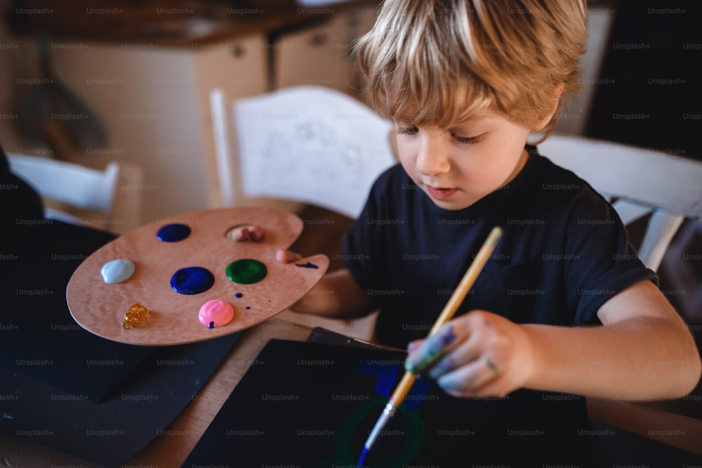 A concentrated small boy painting pictures indoors at home, leisure time